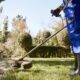 Person in blue coveralls and rubber boots uses a grass trimmer to edge a lawn in a garden with hedges and trees, showcasing the expertise of a Landscape Maintenance Contractor Singapore.